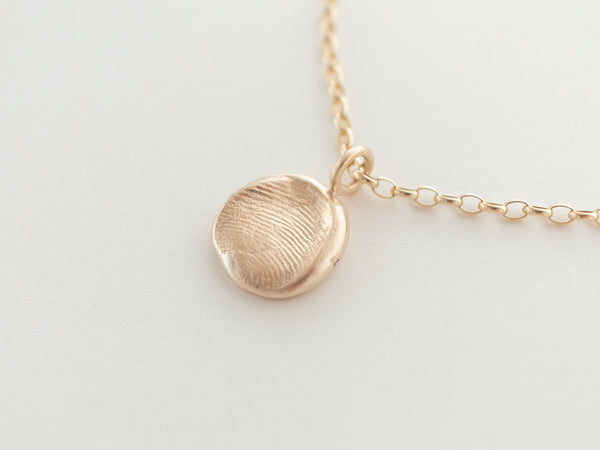 A round gold pendant necklace with a small fingerprint imprint on the front, displayed on a white background.