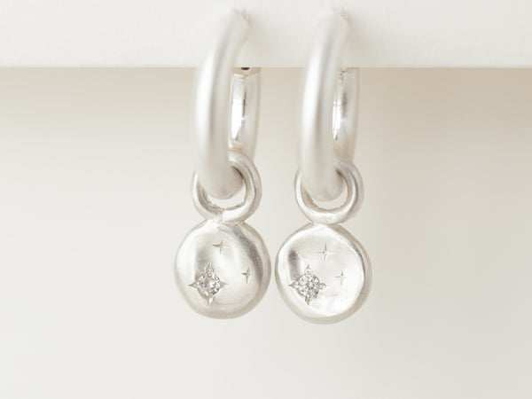 A pair of small silver hoop earrings with a chunky design, featuring two small silver discs with diamonds or moissanites set within, on a white background.