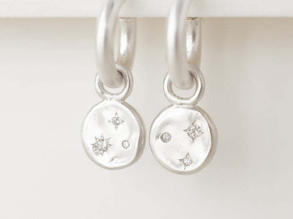 A pair of silver hoop earrings with two small silver discs adorned with diamonds or moissanites, set in hand-engraved stars, displayed against a white background.