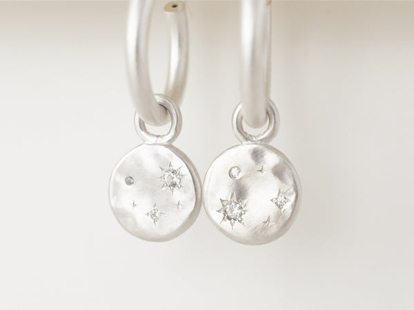 A pair of silver hoop earrings with a starry night design, featuring two silver discs and small diamonds or moissanites set within hand-engraved stars, displayed against a white background.