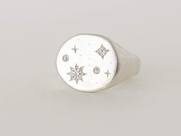 A silver signet ring with a starry night design, featuring a central diamond or moissanite gem surrounded by smaller stones, and engraved details on the band.
