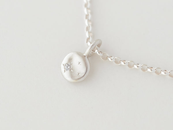 A small silver necklace with a round pendant featuring an offset diamond or moissanite, surrounded by hand-engraved stars, displayed on a white background.