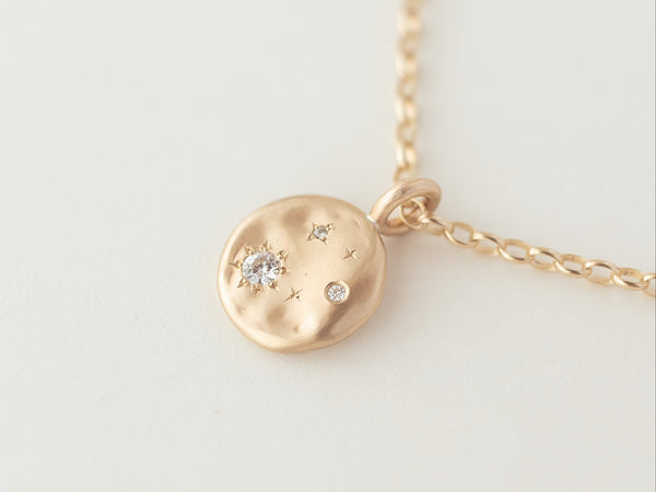A gold necklace featuring a large disc pendant with three Diamonds or Moissanites, surrounded by hand-engraved stars, displayed against a white background.